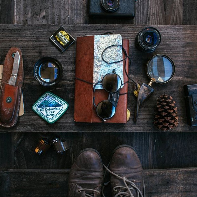 Overhead of vintage photo camera near leather notebook with map and knife near camera roll and lenses near sunglasses placed on table near boots
