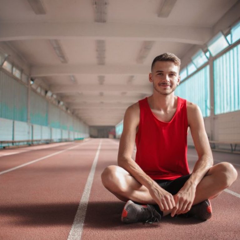 A happy male athlete in a red shirt sitting on an indoor track, relaxing after exercise.