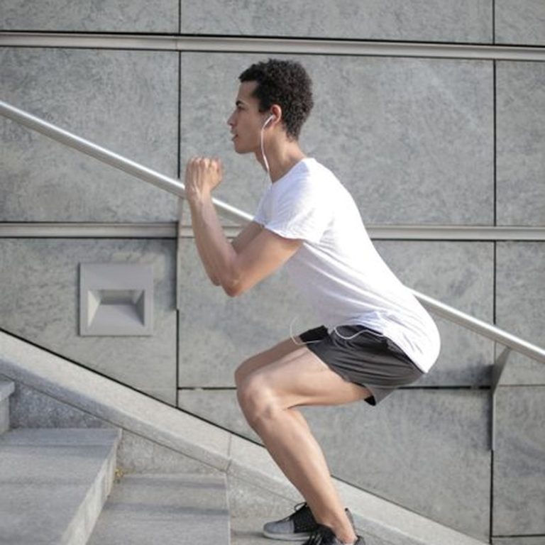 Side view of determined African American man in earphones and sportswear listening to music and jumping on street stairs while training alone against gray wall of modern building