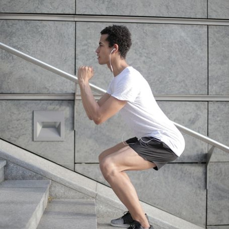Side view of determined African American man in earphones and sportswear listening to music and jumping on street stairs while training alone against gray wall of modern building