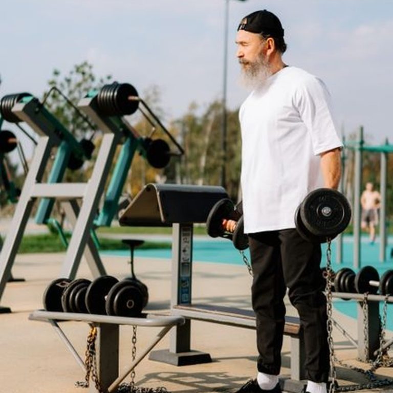 A bearded man in a white shirt works out with dumbbells in an outdoor gym.