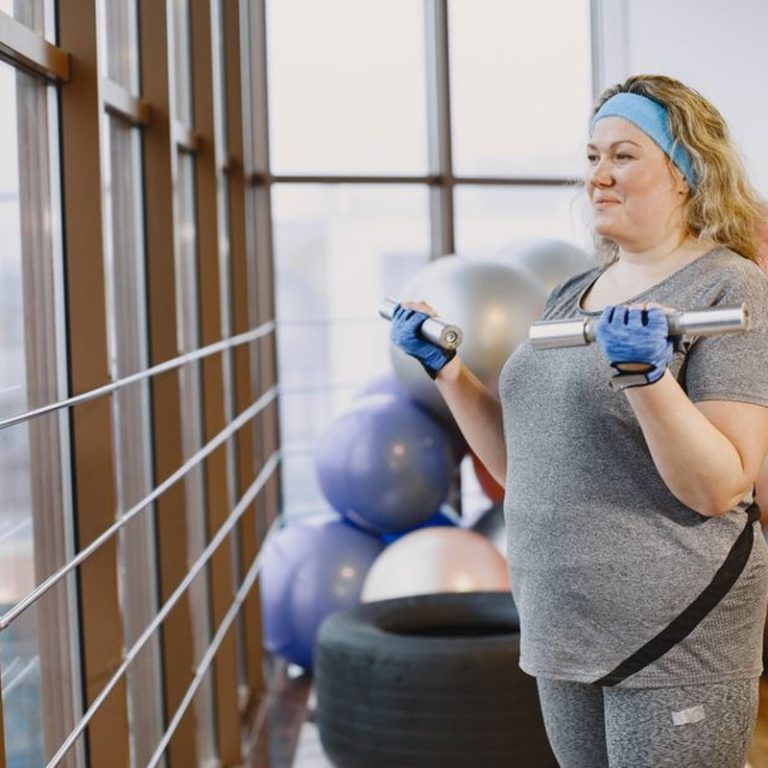 Woman in gym with dumbbells by a window, highlighting fitness and lifestyle.