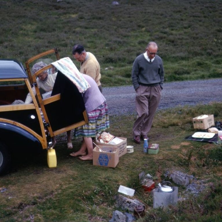 Elderly people enjoying a picnic with a vintage car outdoors.