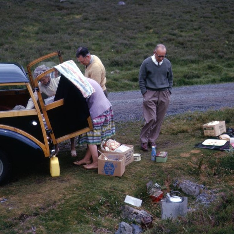 Elderly people enjoying a picnic with a vintage car outdoors.