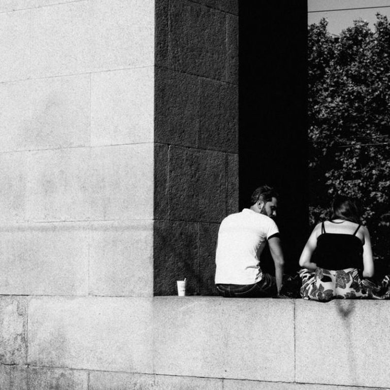 A couple sits together on a concrete ledge outdoors in a black and white setting, facing away.