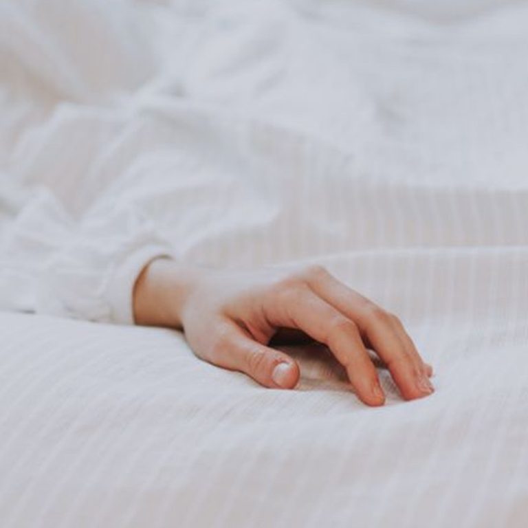 Crop hand of unrecognizable person in white shirt lying on comfortable soft bed in morning