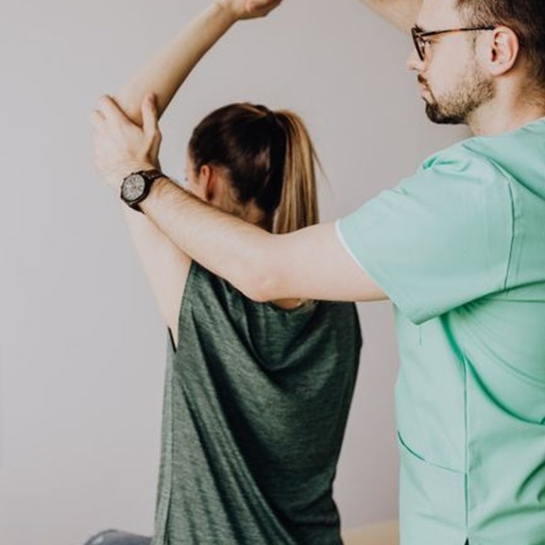 Crop osteopath in eyewear and uniform with wristwatch raising hand of unrecognizable slim woman sitting on examination couch in hospital