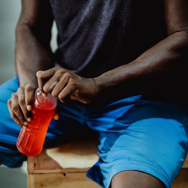 A man resting on a wooden box, enjoying a red sports drink during his workout session.