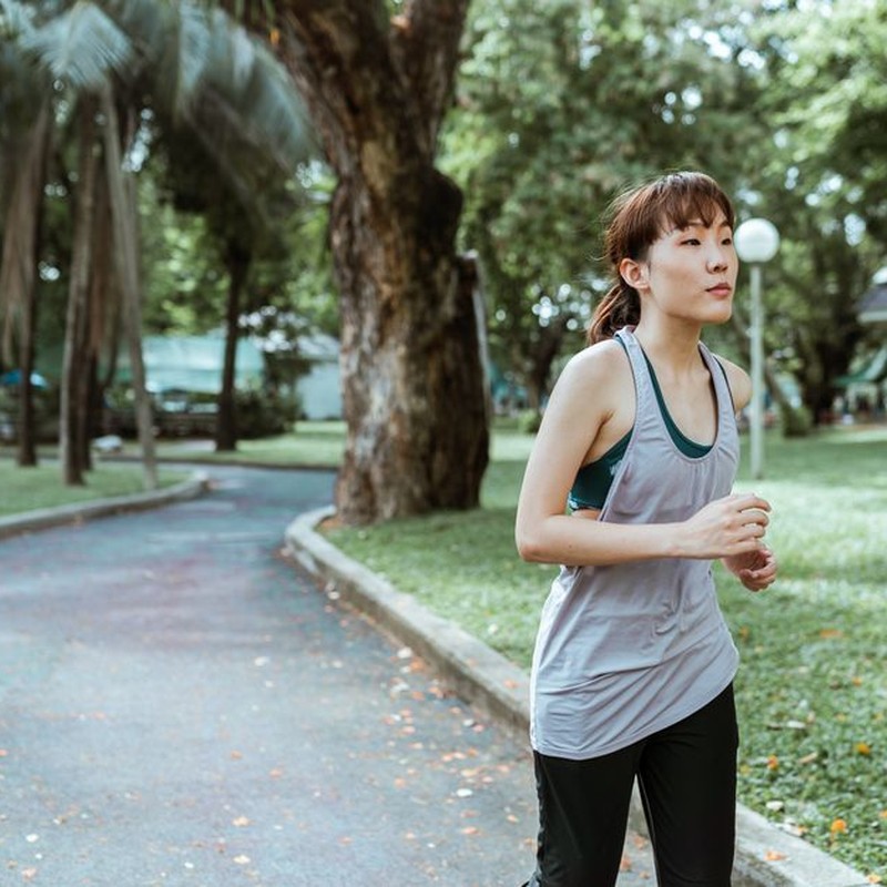 Thoughtful young Asian female athlete in sportswear running on asphalt road covered with faded leaves in city park in daytime and looking away