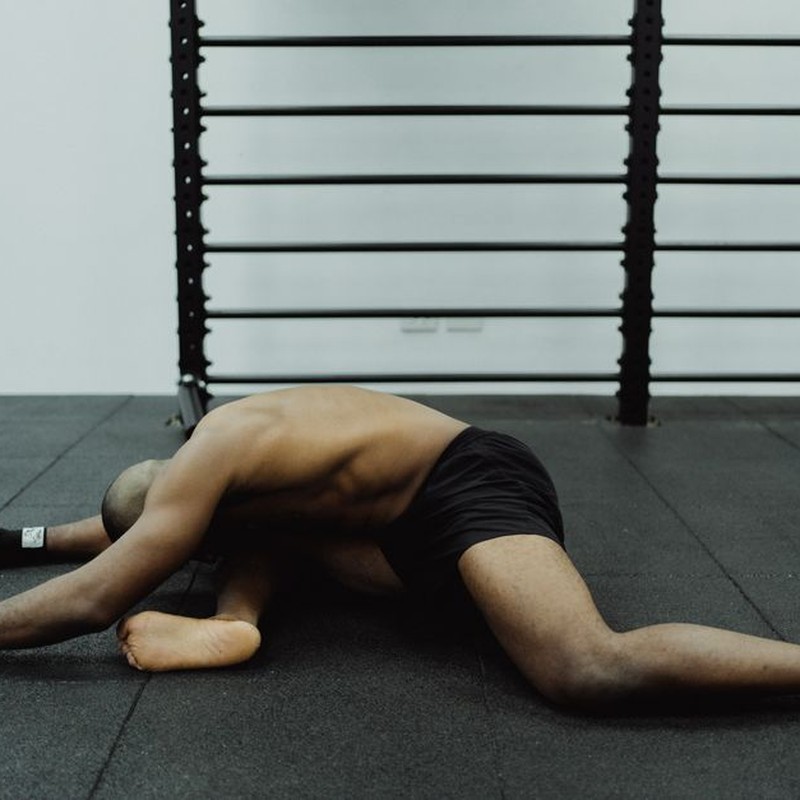 Shirtless man stretches while sitting on gym floor, showcasing flexibility.