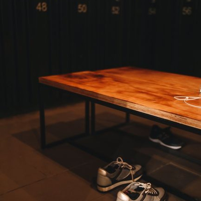 A pair of sneakers under a bench in a locker room with numbered lockers in the background.