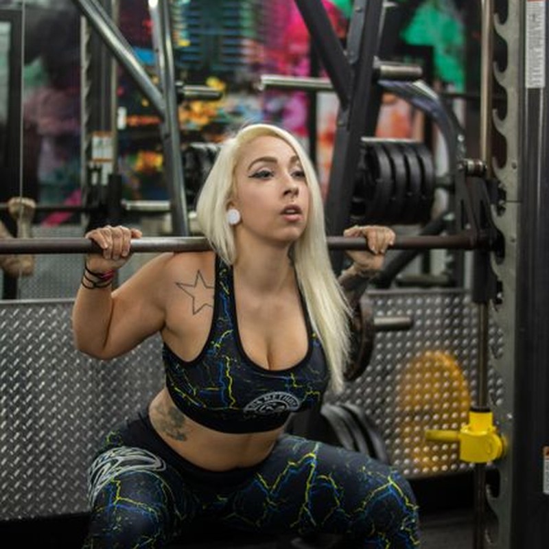 Blonde woman with tattoos working out in a gym using weightlifting equipment.