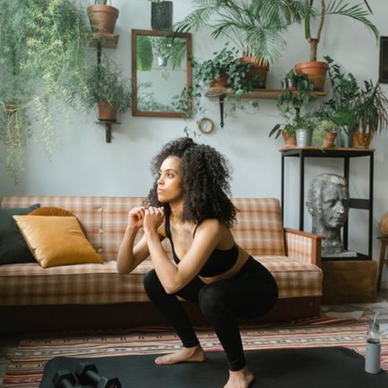 Young woman in active wear doing squat exercises at home with dumbbells and yoga mat.