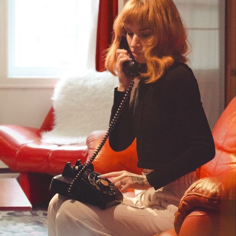 Woman with ginger hair in black long sleeves talks on a vintage phone sitting on a couch indoors.