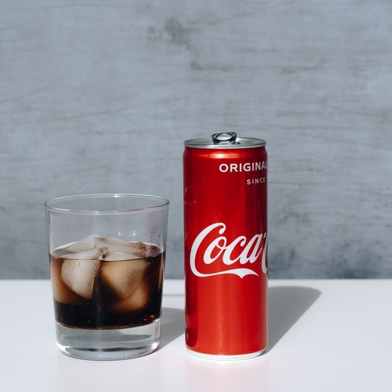 Chilled Coca-Cola can and drink with ice cubes in glass on minimalistic surface.