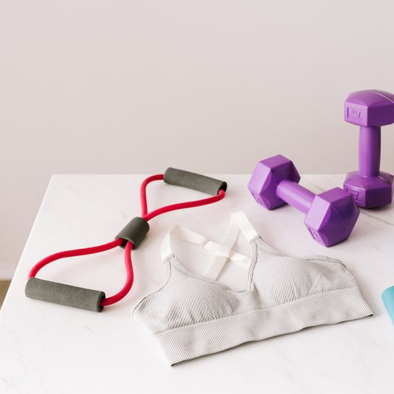 From above of white sports bra surrounded by purple dumbbells and blue tape expander and red tubular expander and blue massage double ball placed on marble surface of table
