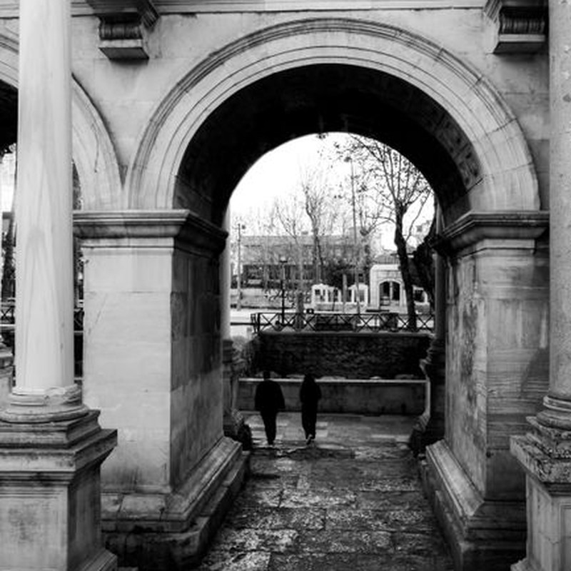 Black and white photo of a classical archway in a city with two people walking through.