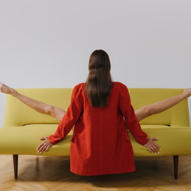 Woman in red blazer sitting creatively on yellow sofa with white wall background.