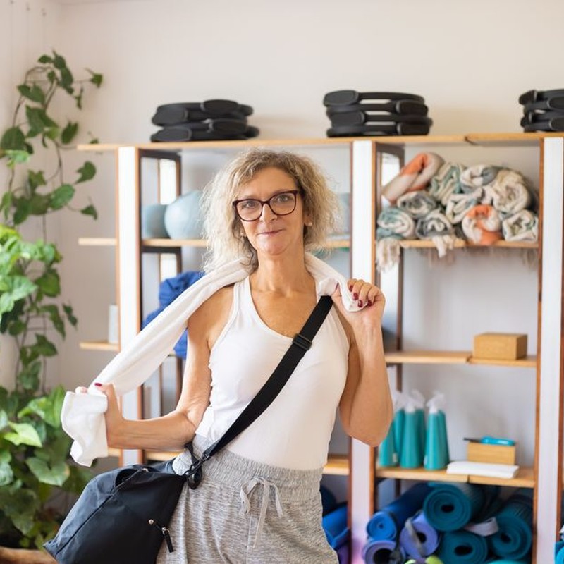 Elderly woman holding a towel in a yoga studio, surrounded by yoga equipment.
