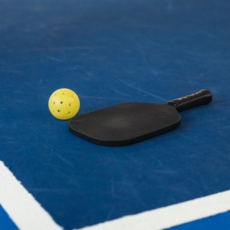 A pickleball paddle and yellow ball on a blue court corner, symbolizing sport and activity.