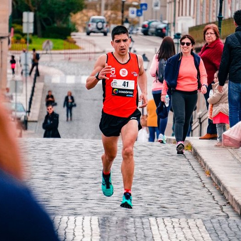 A male athlete runs a marathon through a bustling urban area on a cobblestone street.