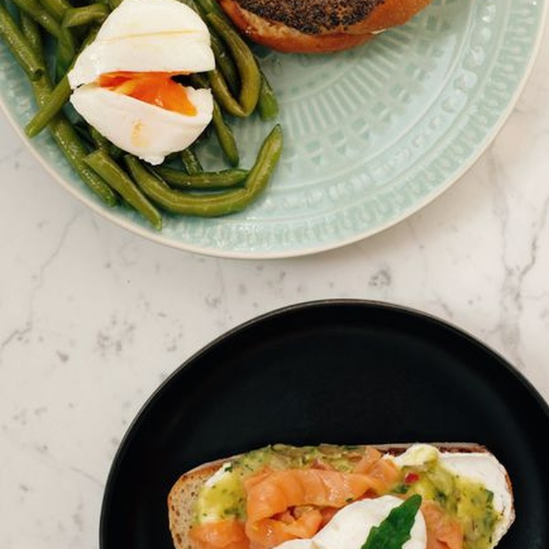 From above of served tasty bagel with asparagus and poached egg and fresh toast with avocado and salmon on white marble table